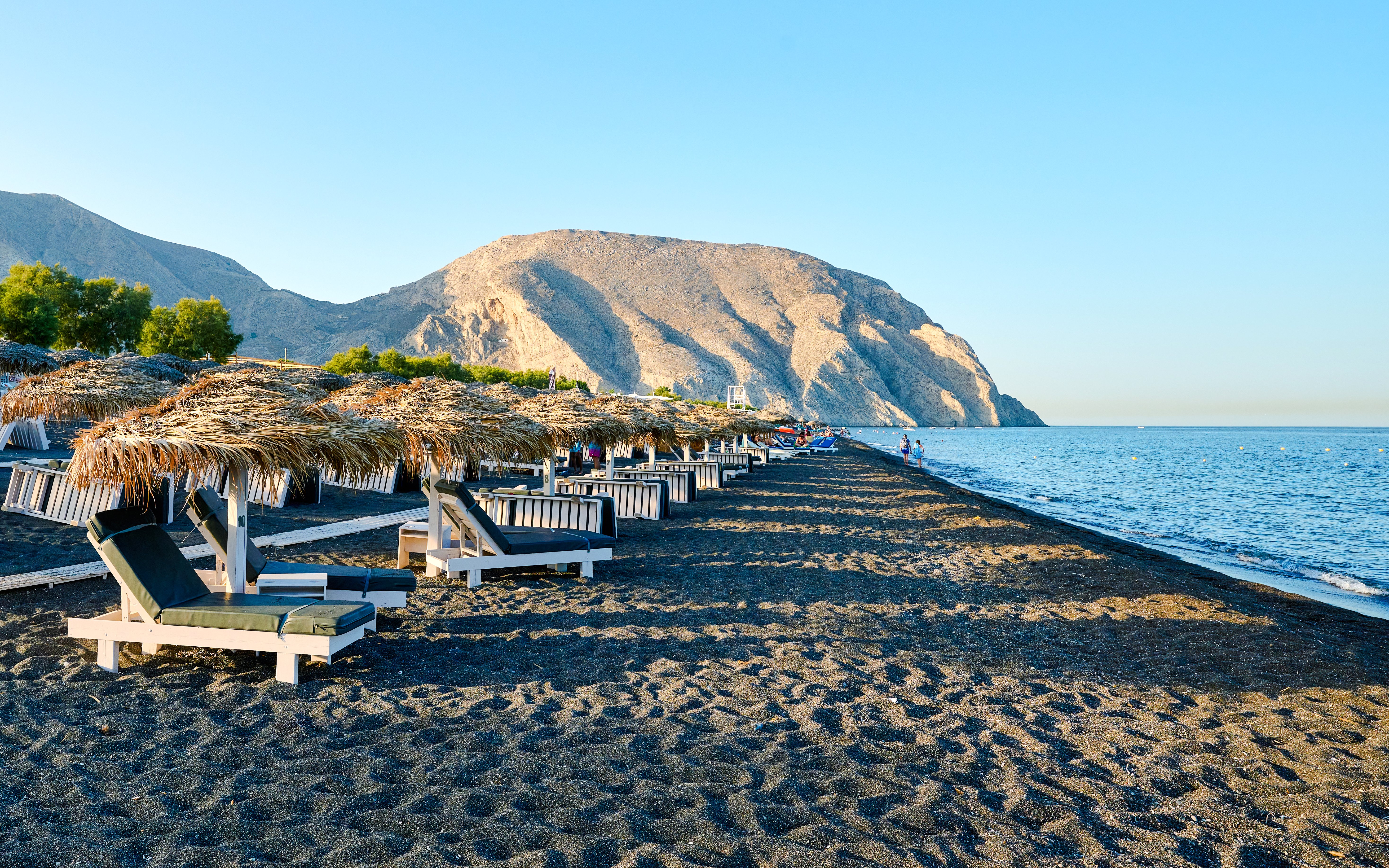 Sun loungers and straw umbrellas on Perivolos Beach, Santorini with mountains in the background.
