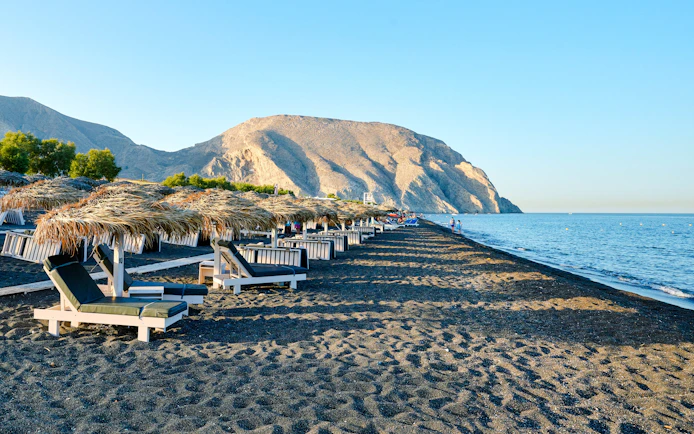 Sun loungers and straw umbrellas on Perivolos Beach, Santorini with mountains in the background.