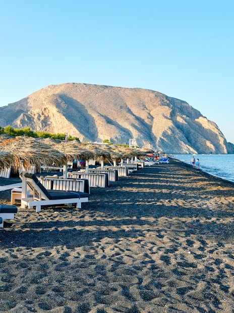 Sun loungers and straw umbrellas on Perivolos Beach, Santorini with mountains in the background.