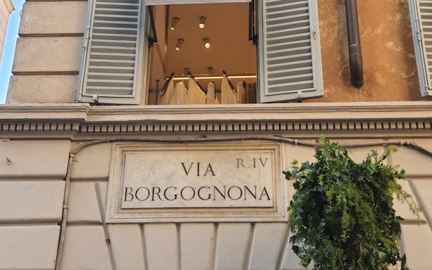 Via Borgognona street sign in Rome, Italy, with open window above displaying fashion garments.