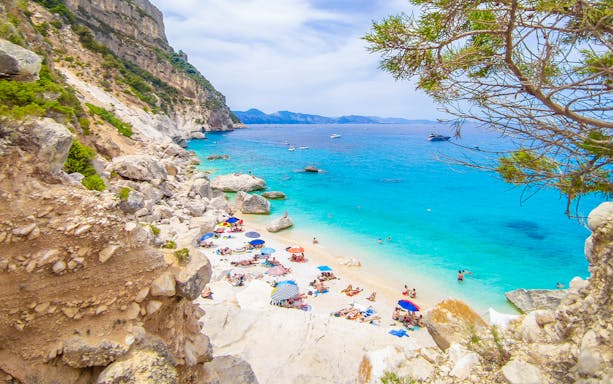 Cala Goloritzè beach in Sardinia with turquoise water, rocky cliffs, and sunbathers on the sand.
