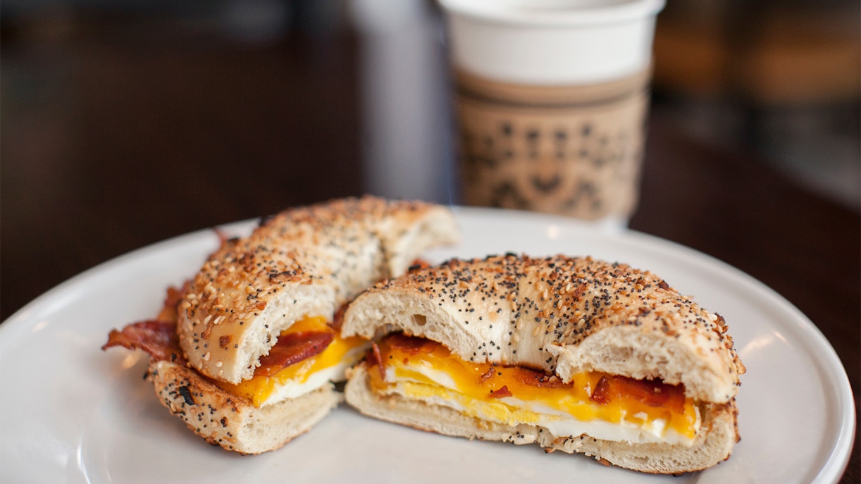 Bagel sandwich with bacon, egg, and cheese on a white plate with a coffee cup in the background.