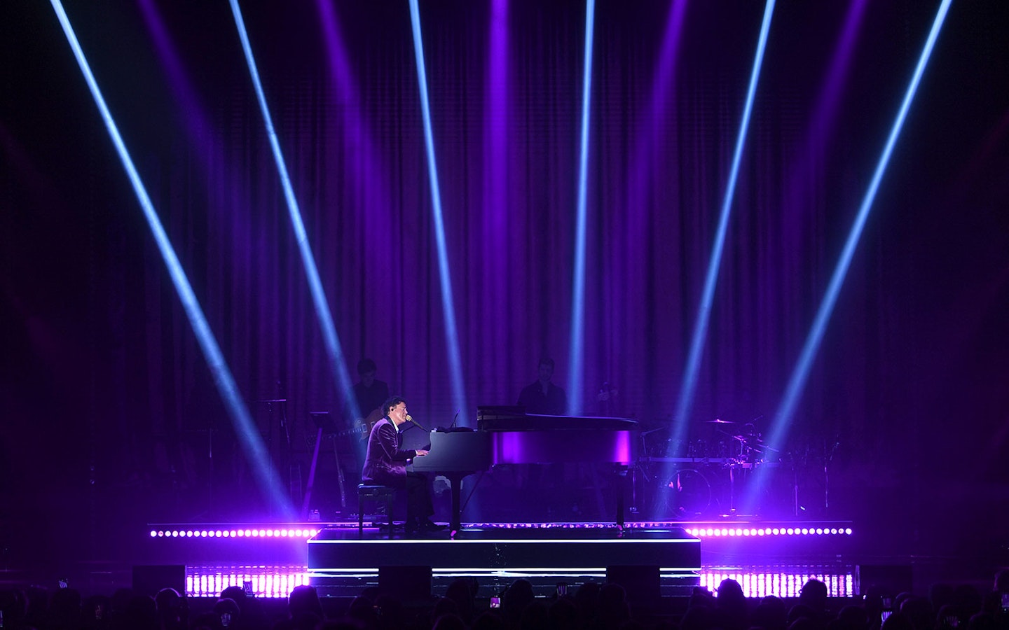 Performer at a grand piano on stage with purple lighting and spotlights.