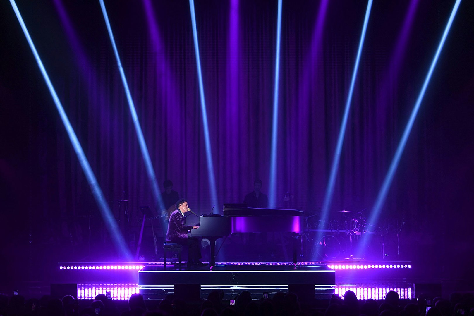 Performer at a grand piano on stage with purple lighting and spotlights.