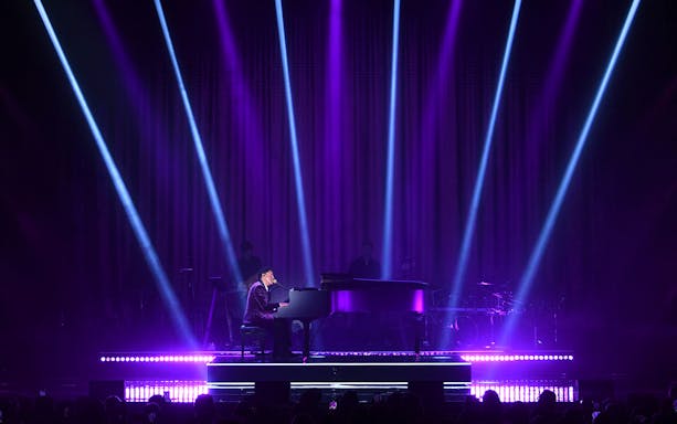 Performer at a grand piano on stage with purple lighting and spotlights.