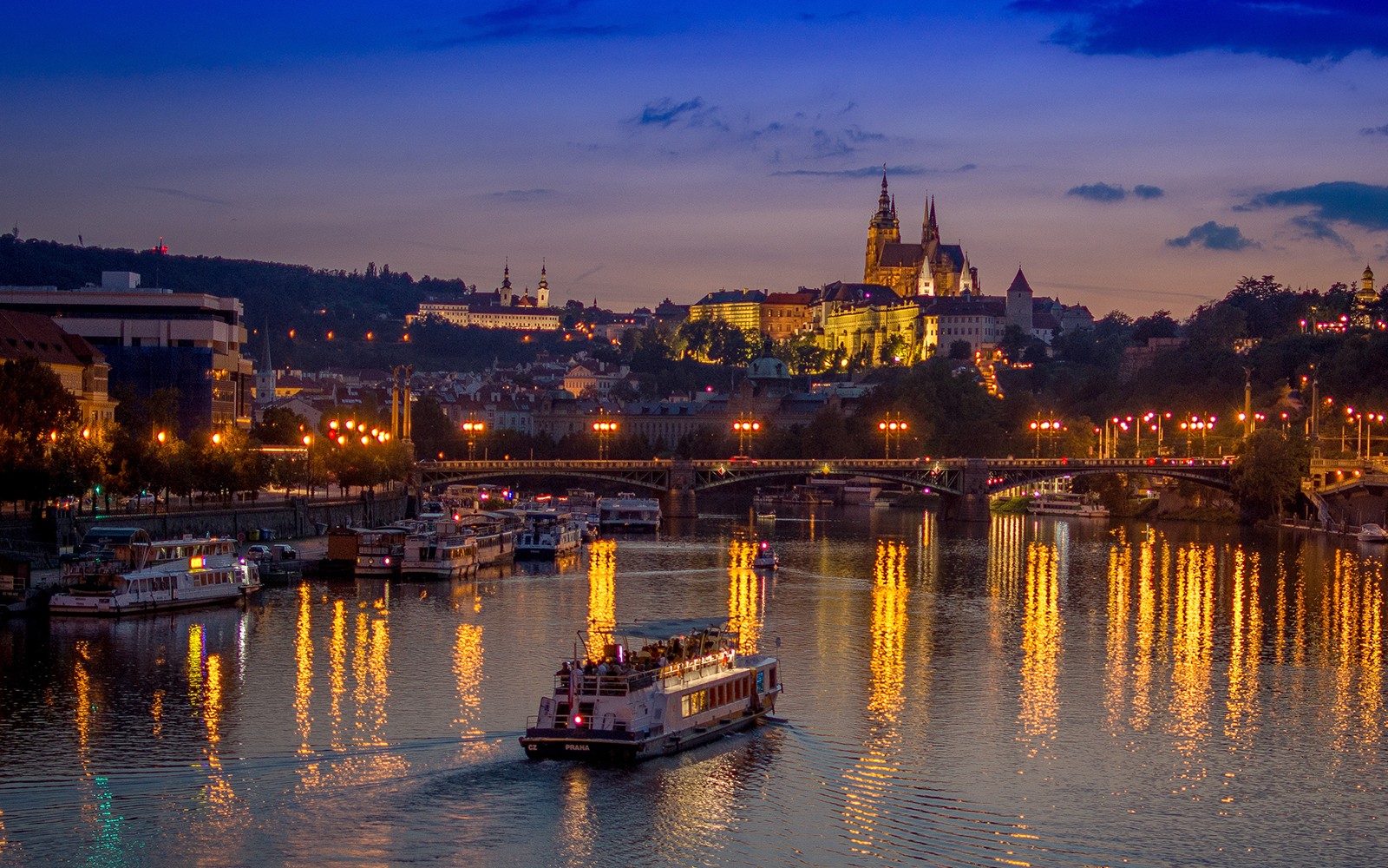 Dinner cruise boat on Vltava River with Prague Castle illuminated at dusk.