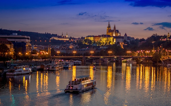 Dinner cruise boat on Vltava River with Prague Castle illuminated at dusk.