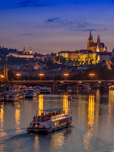 Dinner cruise boat on Vltava River with Prague Castle illuminated at dusk.