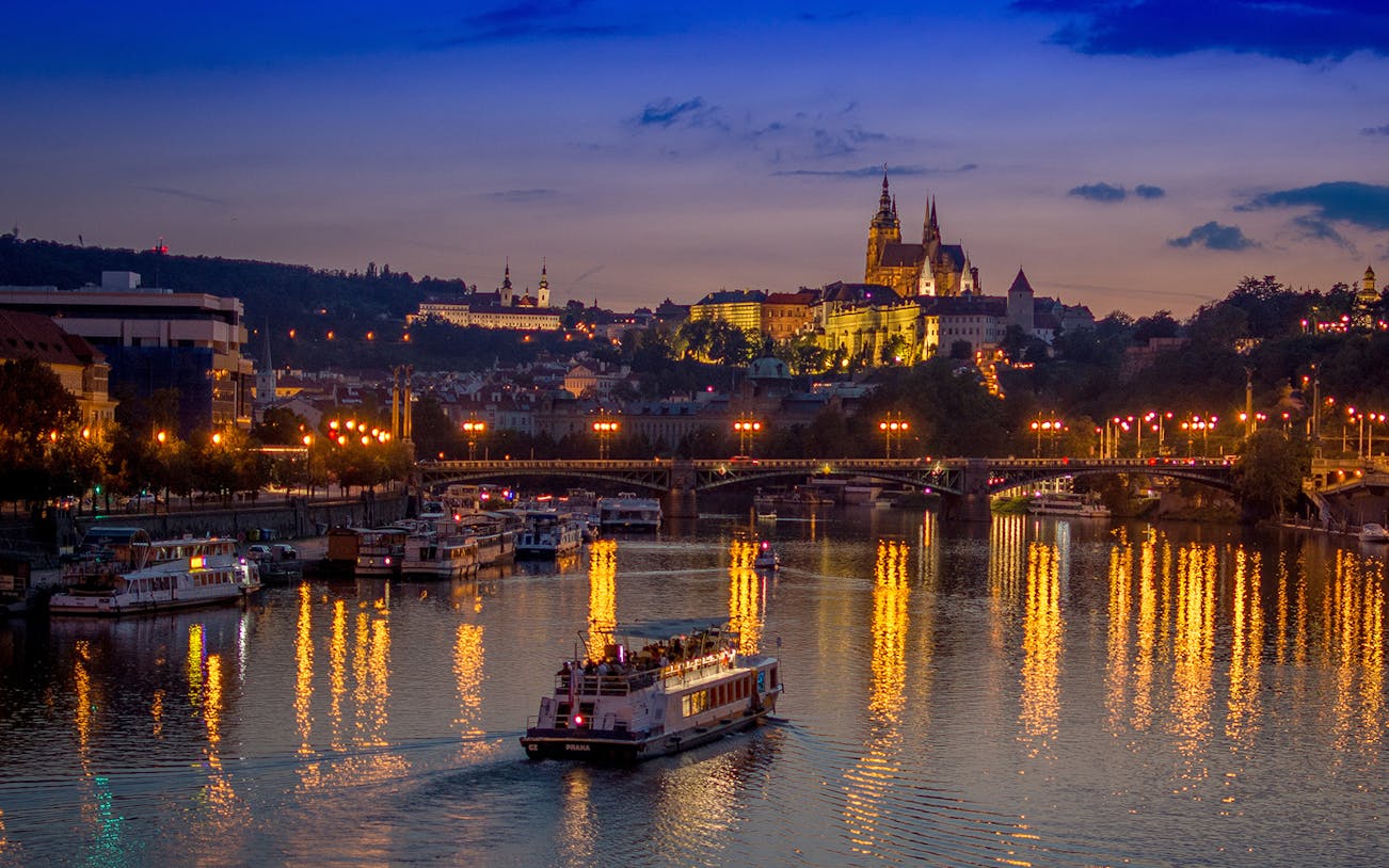 Dinner cruise boat on Vltava River with Prague Castle illuminated at dusk.