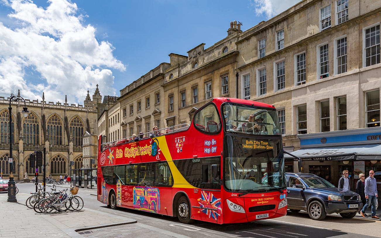 Red double-decker bus on Bath Hop-On Hop-Off Tour passing historic buildings and Bath Abbey.