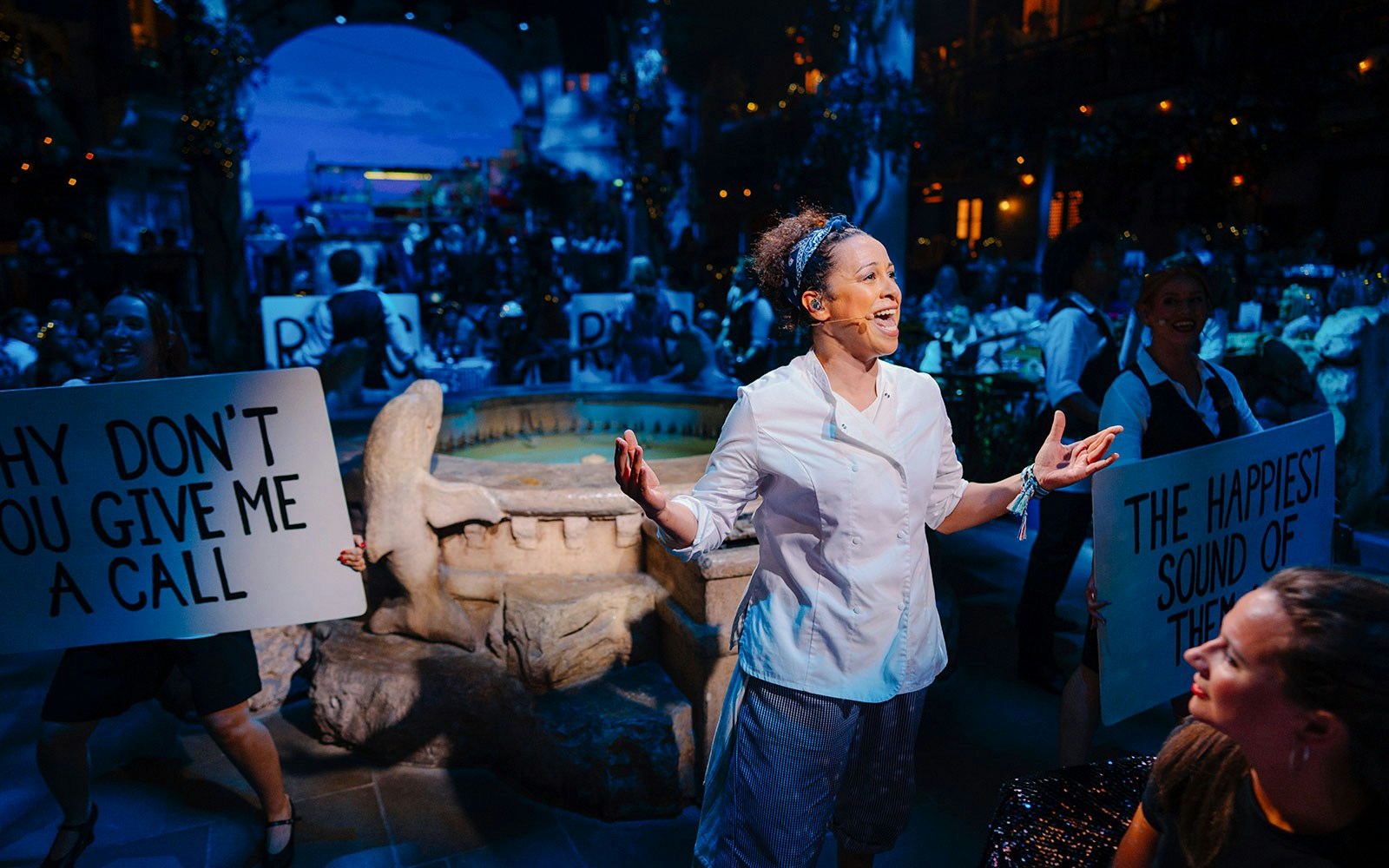 Performer singing at Mamma Mia! The Party with audience holding signs.