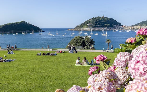 San Sebastian bay view with people relaxing on grass, sailboats in water, and lush greenery.