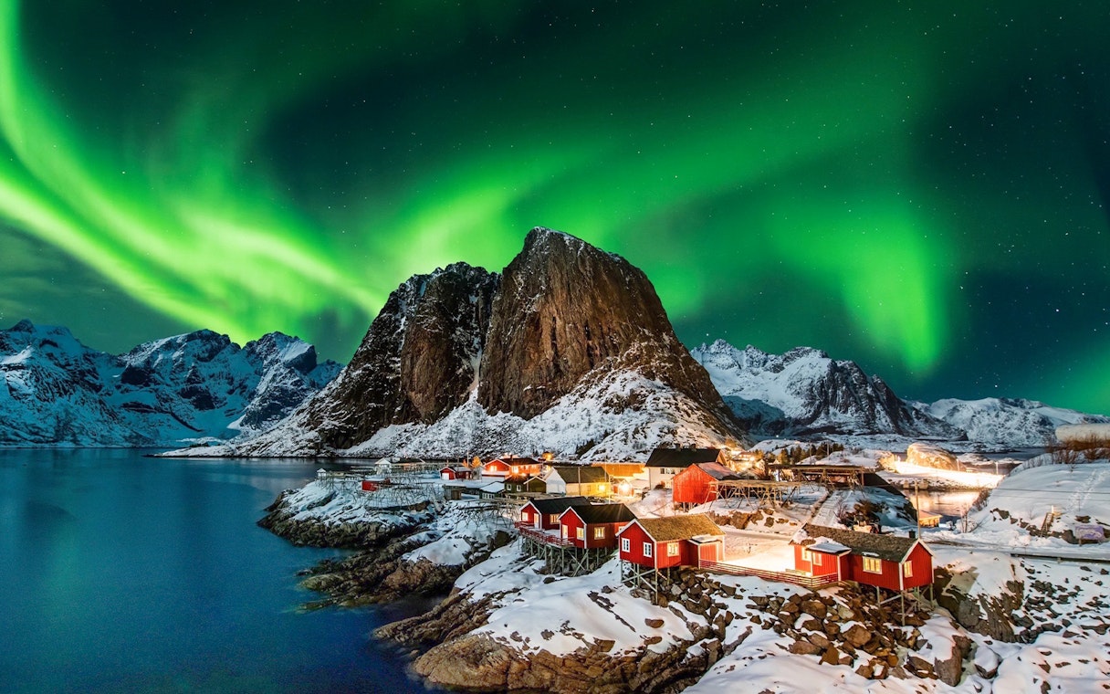 Northern lights over snowy mountains and red cabins in Lofoten, Norway.