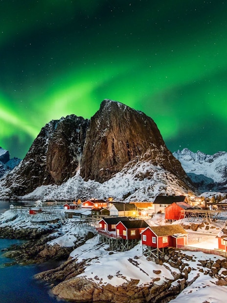 Northern lights over snowy mountains and red cabins in Lofoten, Norway.