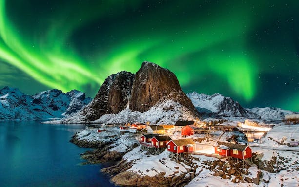 Northern lights over snowy mountains and red cabins in Lofoten, Norway.