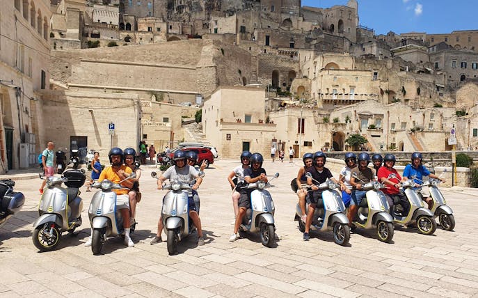 Group on e-Vespas touring Sassi di Matera, Italy, with historic stone buildings in the background.