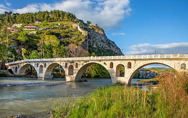 Historical arched bridge spanning the Osumi River in Berat, Albania, with scenic mountainous backdrop.