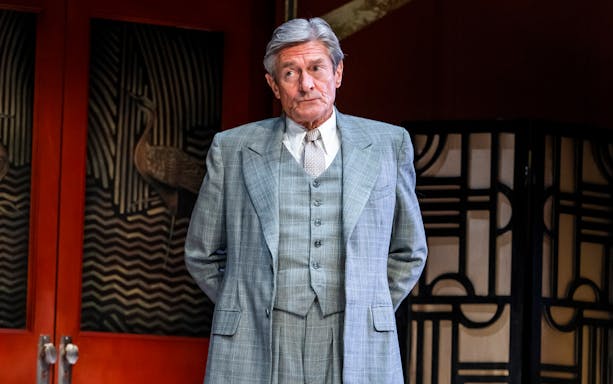 Man in a gray suit standing in front of an art deco backdrop during "Private Lives" performance.