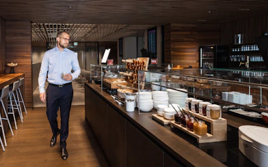 Man walking in BER Lounge Tegel at Berlin Brandenburg Airport, near buffet with pretzels and condiments.