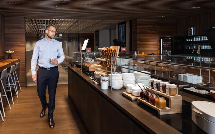 Man walking in BER Lounge Tegel at Berlin Brandenburg Airport, near buffet with pretzels and condiments.