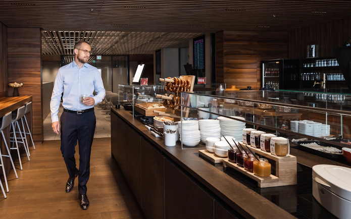 Man walking in BER Lounge Tegel at Berlin Brandenburg Airport, near buffet with pretzels and condiments.