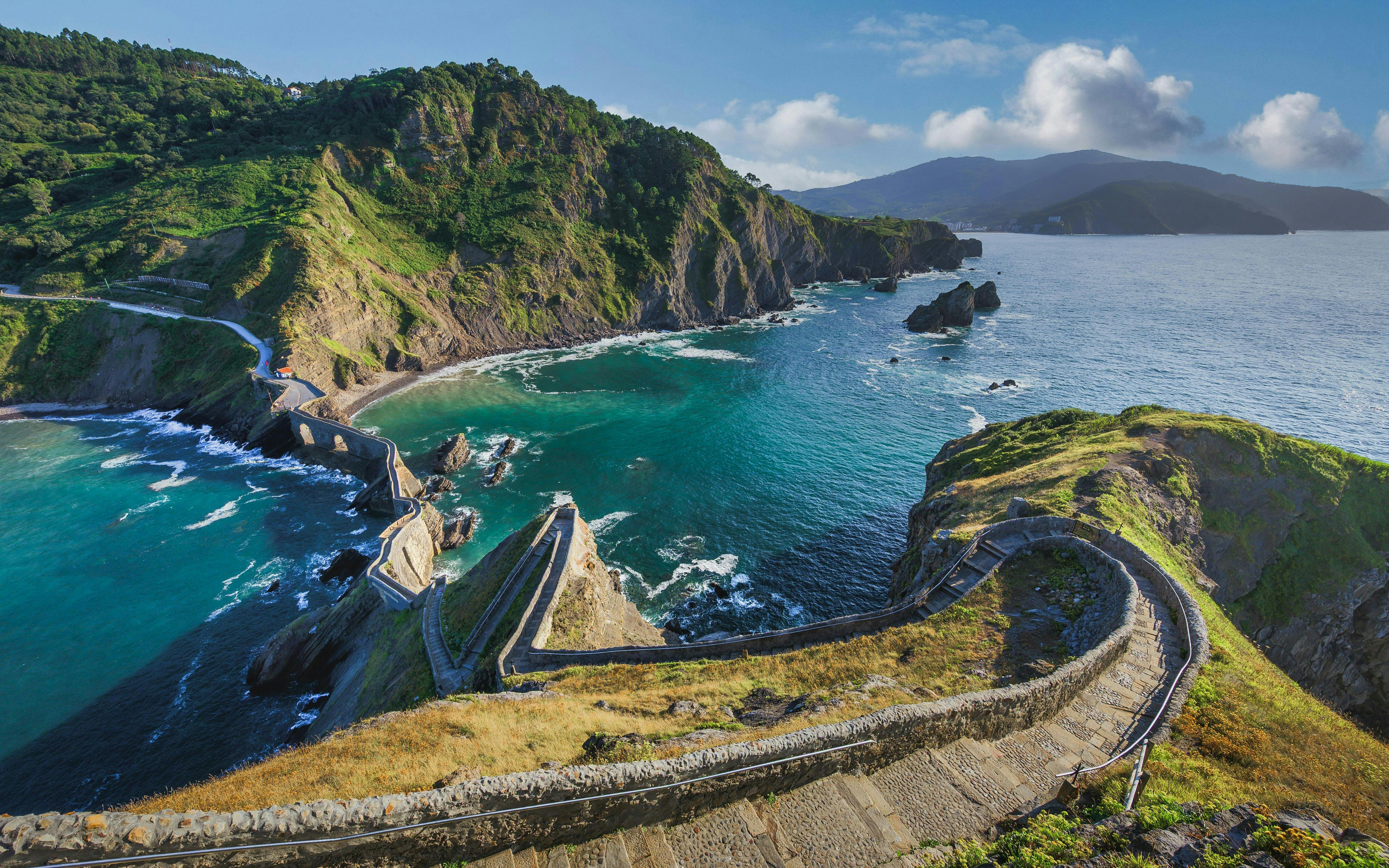 Winding stone staircase descending to coastal bay with turquoise waters and rocky cliffs, San Juan de Gaztelugatxe, Spain.