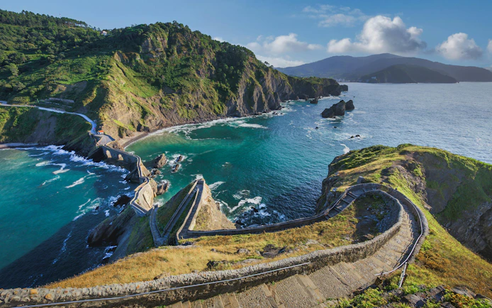 Winding stone staircase descending to coastal bay with turquoise waters and rocky cliffs, San Juan de Gaztelugatxe, Spain.