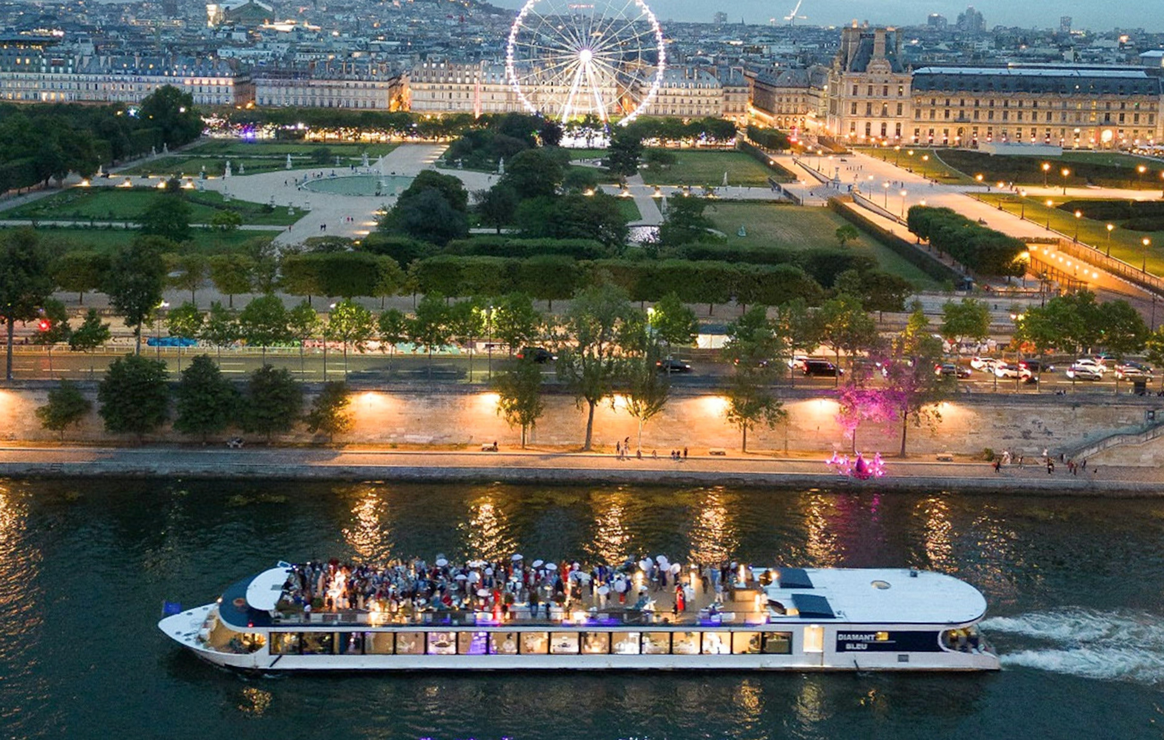 Seine River dinner cruise boat with guests, Paris Ferris wheel and city lights in background.
