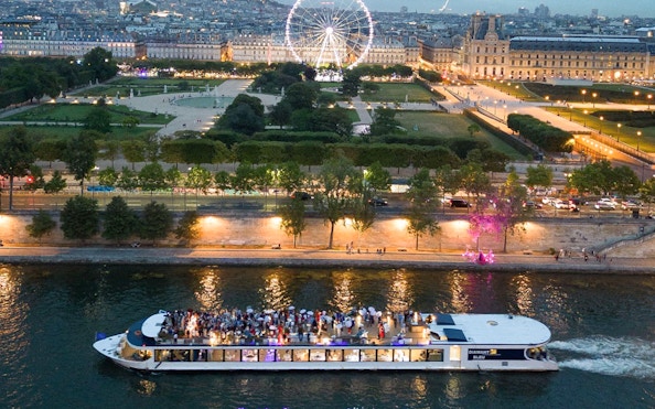 Seine River dinner cruise boat with guests, Paris Ferris wheel and city lights in background.