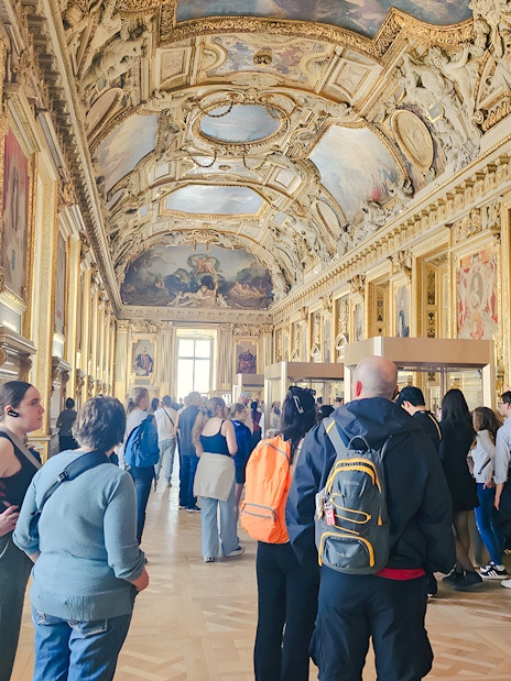 Visitors exploring ornate gallery inside Louvre Museum, Paris, France.