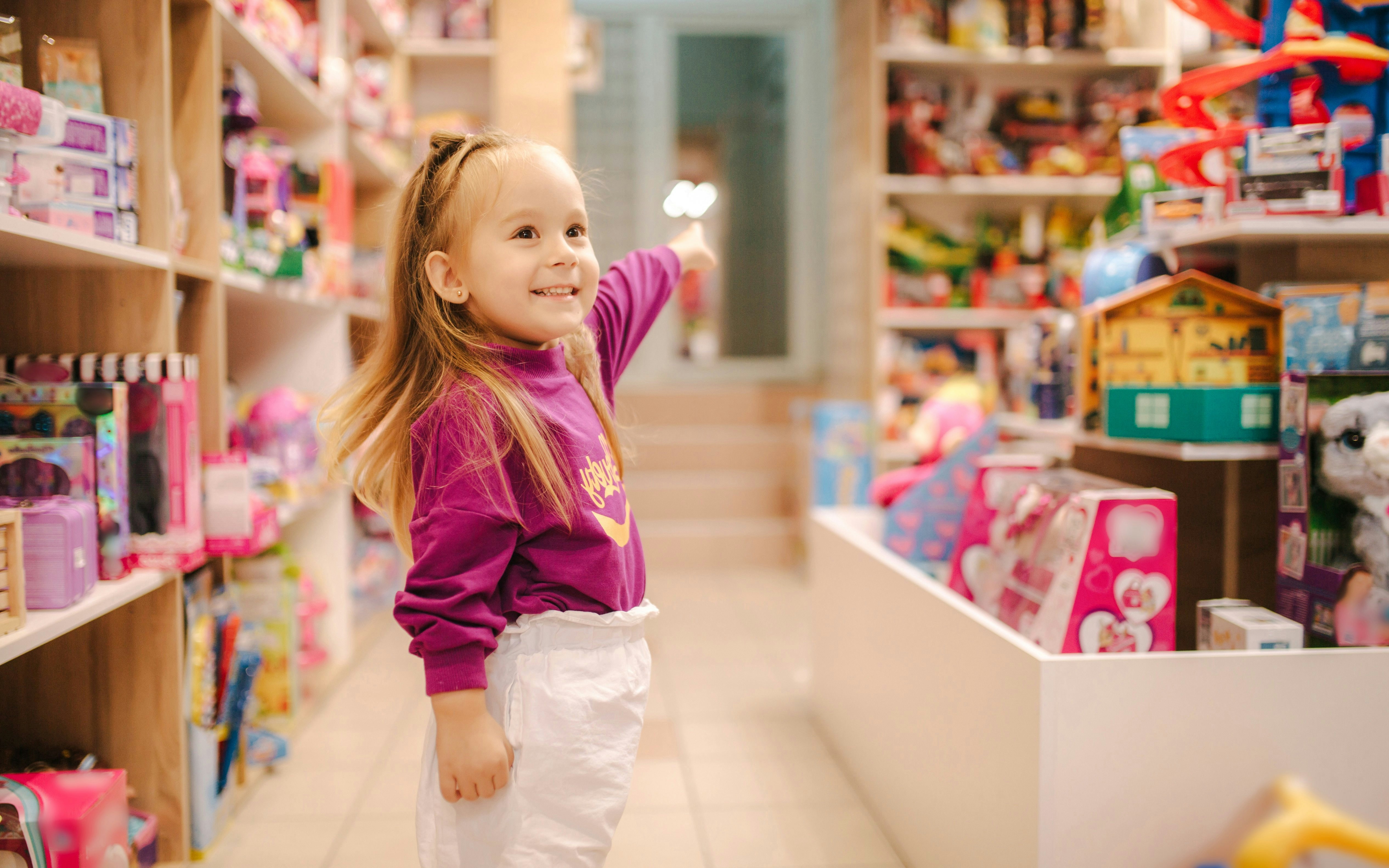Little girl pointing at toys in a colorful toy store aisle.