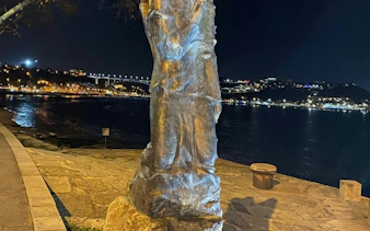 Statue by the Douro River at night in Porto with city lights in the background.