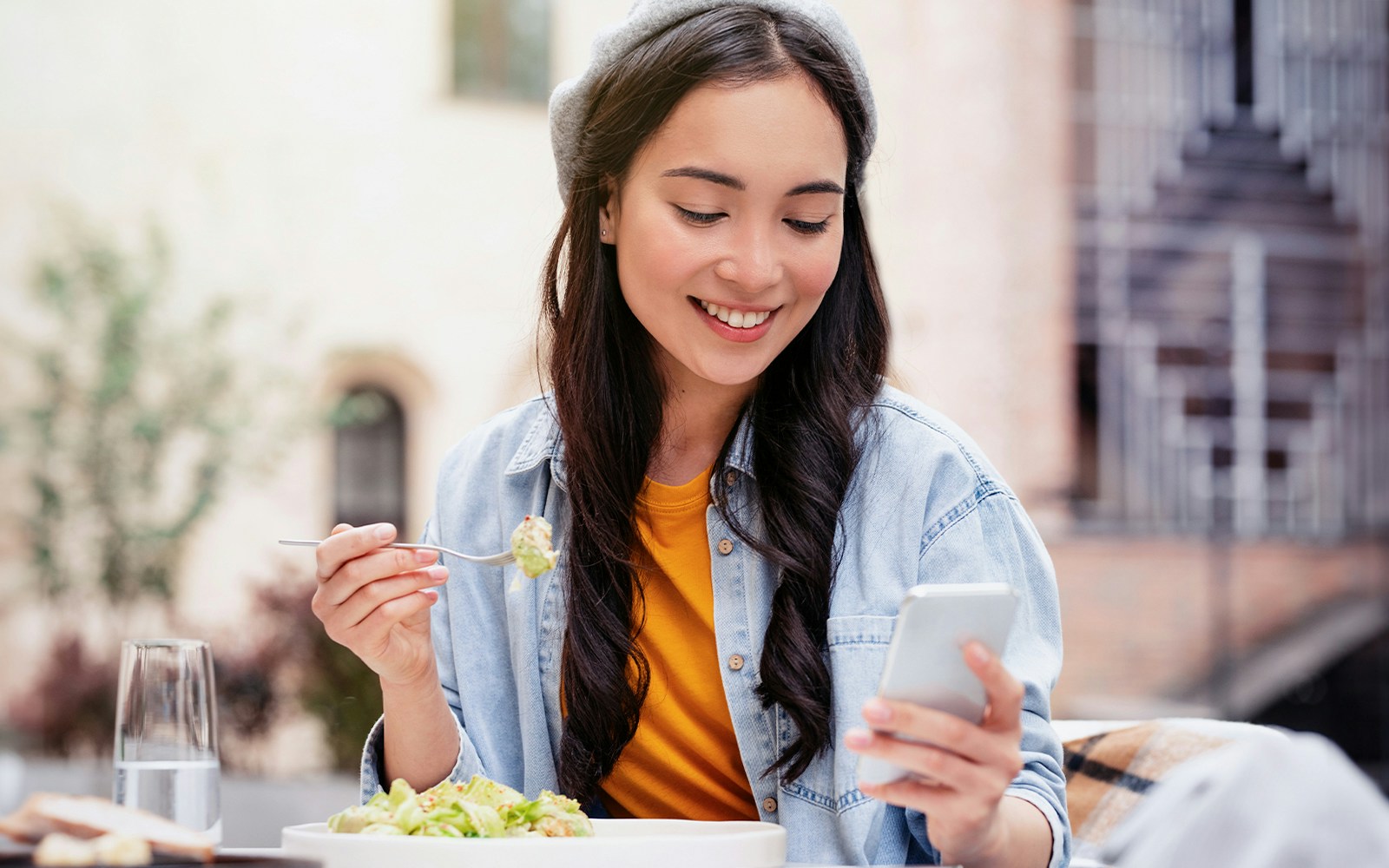 Woman using smartphone while eating salad at outdoor cafe.