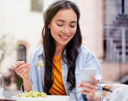 Woman using smartphone while eating salad at outdoor cafe.