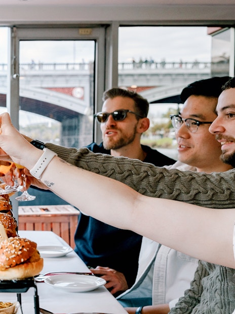 Guests toasting with drinks on a brunch cruise in Melbourne, with burgers on the table.