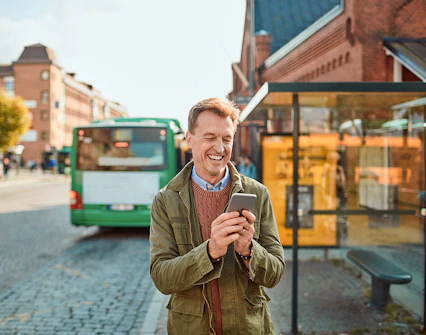 Person smiling while using phone at a bus stop with a green bus in the background.