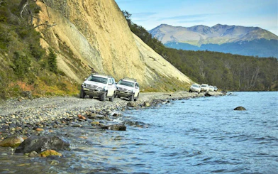 Off-road 4x4 vehicles driving along the shore of Lake Fagnano, surrounded by mountains and forest.