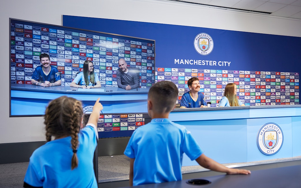 Children experiencing a press conference setup during a guided tour of Manchester City Stadium.
