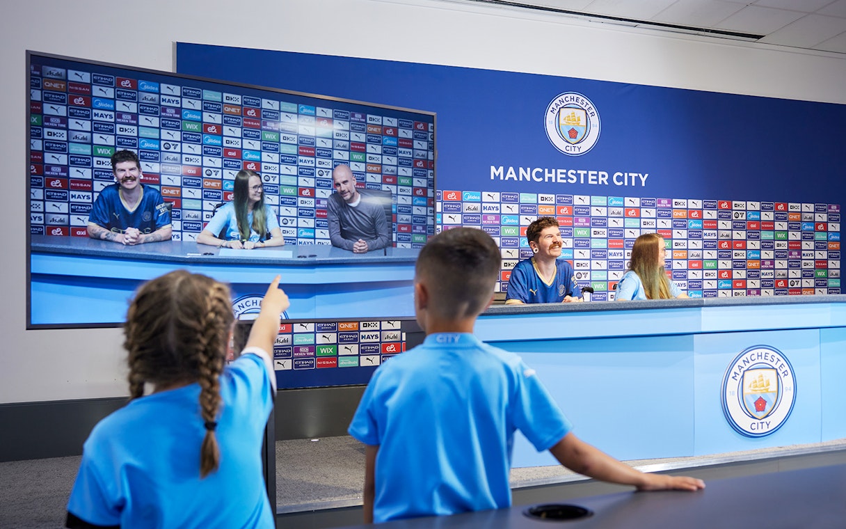 Children experiencing a press conference setup during a guided tour of Manchester City Stadium.