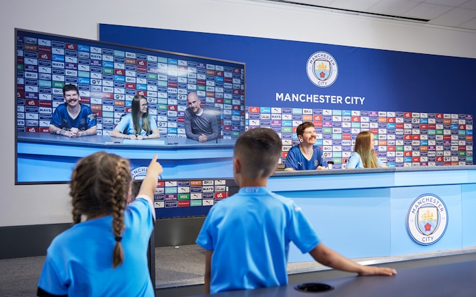Children experiencing a press conference setup during a guided tour of Manchester City Stadium.