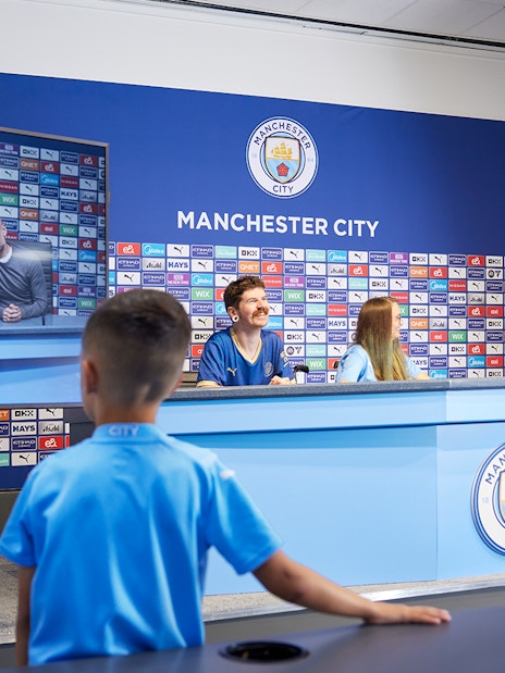 Children experiencing a press conference setup during a guided tour of Manchester City Stadium.