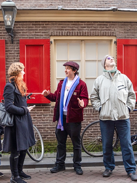 Tour guide leading a group in Amsterdam's Red Light District, standing by red shuttered windows.