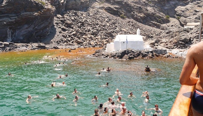 Guests swimming in hot springs near a white chapel during Santorini volcanic cruise tour.
