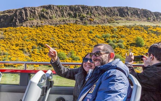 Tourists enjoying a Hop on Hop off bus tour with scenic yellow flowers and rocky hills in the background.