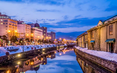 Otaru Canal in Hokkaido during winter evening with snow-covered banks and illuminated buildings.