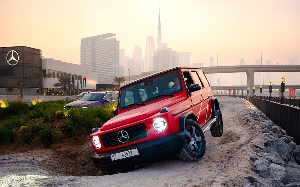 Red Mercedes-Benz G-Class on off-road track in Dubai with city skyline in background.