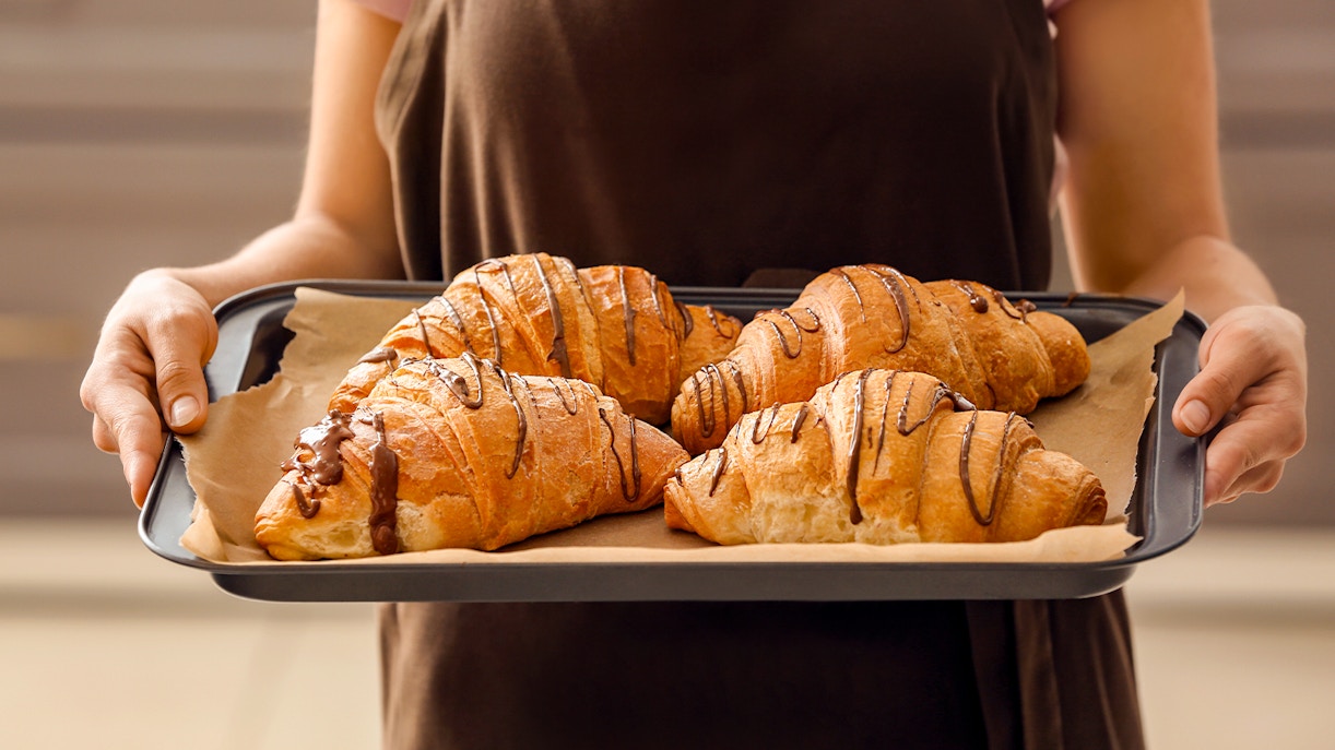 Young woman holding tray of freshly baked croissants in bakery.