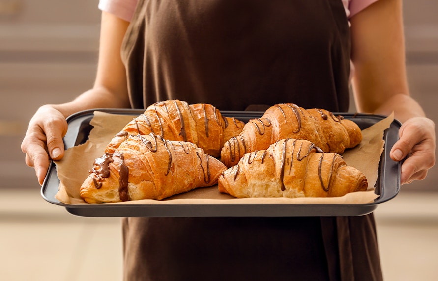 Young woman holding tray of freshly baked croissants in bakery.
