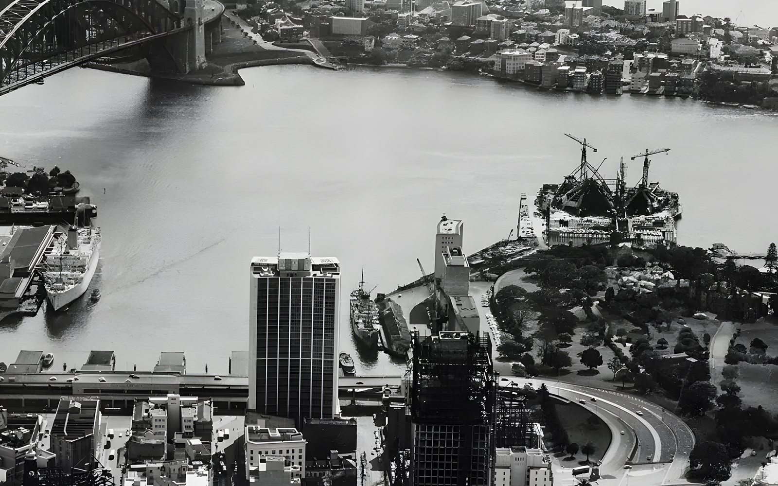 Sydney Opera House under construction with cranes, view of Sydney Harbour Bridge in the background.