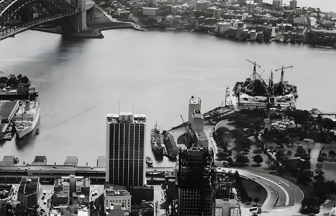 Sydney Opera House under construction with cranes, view of Sydney Harbour Bridge in the background.
